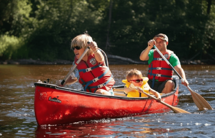 Three people, two adults and a child, wearing life jackets and paddling a red canoe on a calm river surrounded by trees.