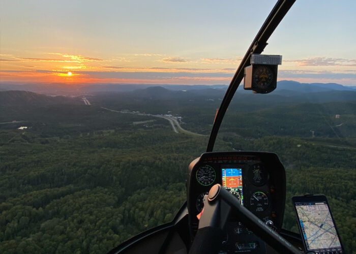 View from inside a helicopter cockpit flying over a forested landscape at sunset, with flight control panels and a navigation screen visible.