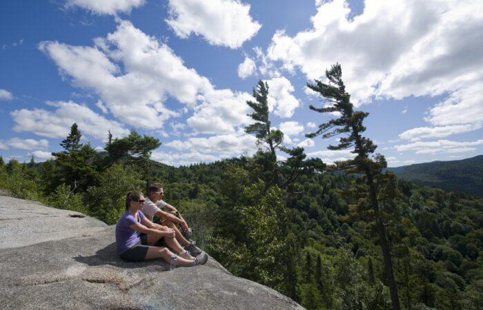 Deux personnes sont assises sur une corniche rocheuse surplombant une vallée boisée et des collines sous un ciel partiellement nuageux.