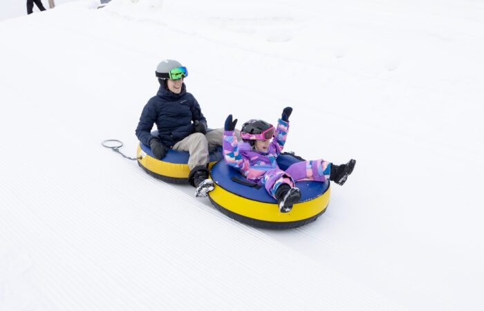 Un adulte et un enfant portant des vêtements d'hiver dévalent une colline enneigée sur des tubes de neige jaunes et bleus.