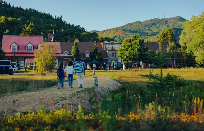 Three people walk on a dirt path toward a lodge surrounded by trees and mountains on a clear day. Three people walk on a dirt path toward a lodge surrounded by trees and mountains on a clear day.