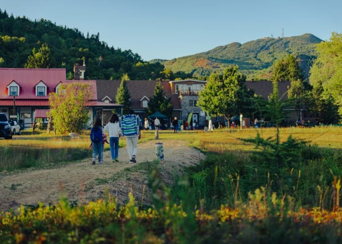 Three people walk on a dirt path toward a lodge surrounded by trees and mountains on a clear day.