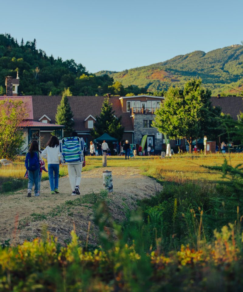 Three people walk on a dirt path toward a lodge surrounded by trees and mountains on a clear day.