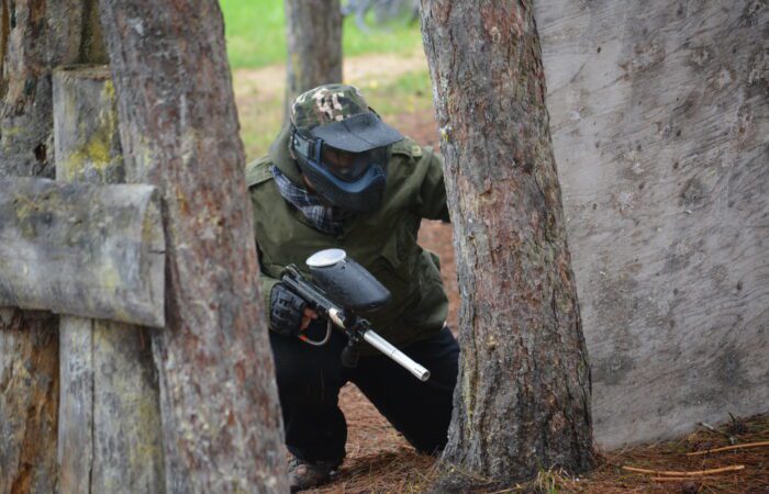 Une personne portant un équipement de protection et tenant un pistolet de paintball est accroupie derrière des arbres et une barrière en bois sur un terrain de paintball en plein air.