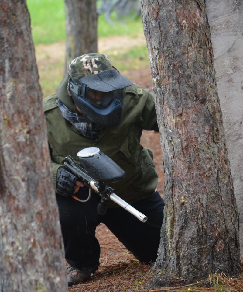 Une personne portant un équipement de protection et tenant un pistolet de paintball est accroupie derrière des arbres et une barrière en bois sur un terrain de paintball en plein air.