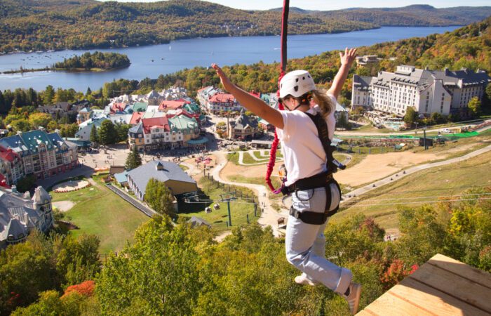 A person wearing safety gear jumps from a platform on a zipline above a resort village with a lake and forested hills in the background, where adventure seekers can also enjoy Guided 4×4 Tours through scenic trails.
