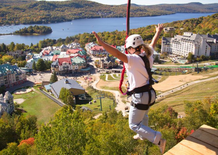 A person wearing safety gear jumps from a platform on a zipline above a resort village with a lake and forested hills in the background, where adventure seekers can also enjoy Guided 4×4 Tours through scenic trails.