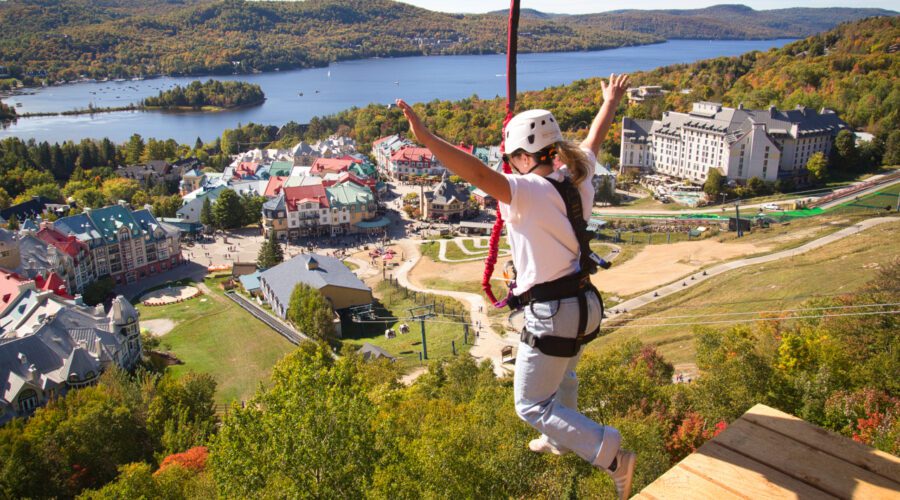 A person wearing safety gear jumps from a platform on a zipline above a resort village with a lake and forested hills in the background, where adventure seekers can also enjoy Guided 4×4 Tours through scenic trails. A person wearing safety gear jumps from a platform on a zipline above a resort village with a lake and forested hills in the background, where adventure seekers can also enjoy Guided 4×4 Tours through scenic trails.