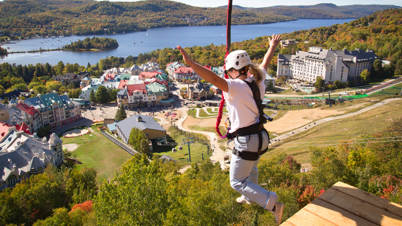 A person wearing safety gear jumps from a platform on a zipline above a resort village with a lake and forested hills in the background, where adventure seekers can also enjoy Guided 4×4 Tours through scenic trails.