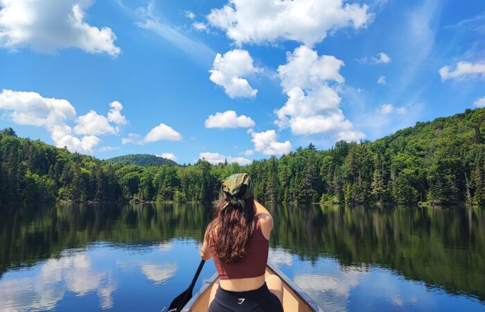 A person paddles a canoe on a calm lake surrounded by green forest under a bright blue sky with scattered clouds, enjoying one of the classic low-impact activities popular in Mont-Tremblant during summer and fall.