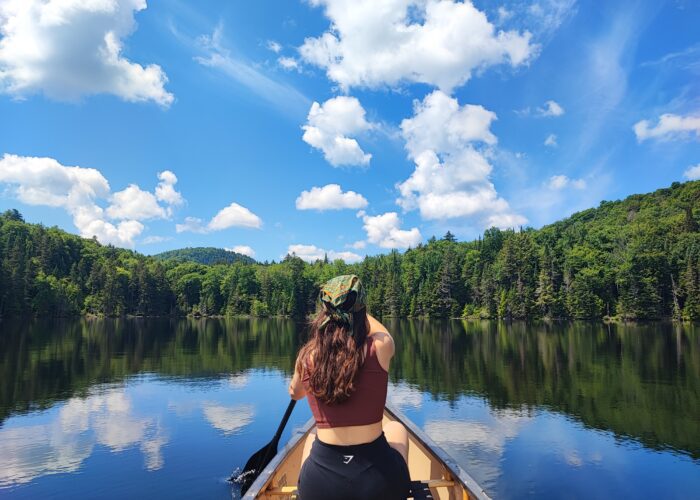 A person paddles a canoe on a calm lake surrounded by green forest under a bright blue sky with scattered clouds, enjoying one of the classic low-impact activities popular in Mont-Tremblant during summer and fall. A person paddles a canoe on a calm lake surrounded by green forest under a bright blue sky with scattered clouds, enjoying one of the classic low-impact activities popular in Mont-Tremblant during summer and fall.