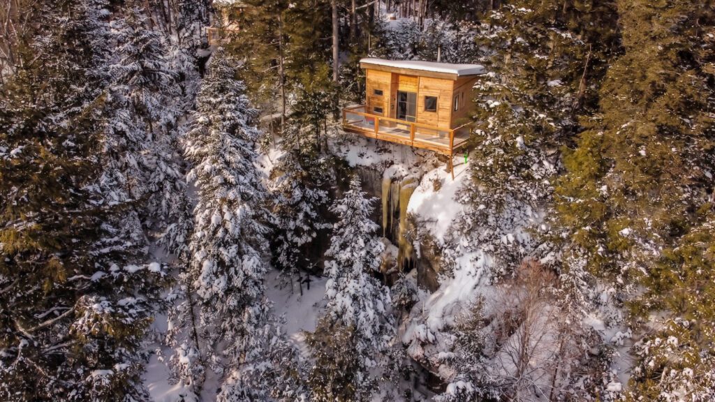 A wooden cabin with a deck sits on the edge of a snowy cliff surrounded by pine trees, overlooking a small waterfall in a winter forest—an ideal Mont-Tremblant retreat for enjoying low-impact activities and embracing the peaceful winter outdoors.